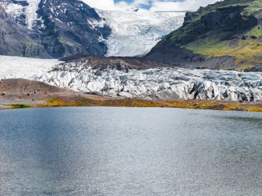 İzlanda 'daki bir buzul engebeli uçurumlar, volkanik kayalar, yeşil yosunlar ve parçalı bulutlu bir gökyüzü tarafından çerçevelenmiş sakin bir göle akar..