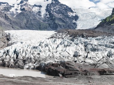 Tırtıklı buzullar, volkanik kayalar ve çamurlu bir buzulla çevrili engebeli kar örtülü dağlardan uzanır. Bir kişi sahneye ölçek ekler.