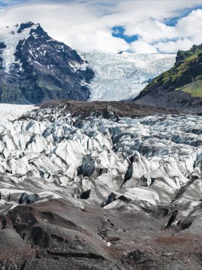 İzlanda 'da buz kütleli bir buzul, karla kaplı bir dağ ve parçalı bulutlu bir gökyüzünün altında yosun kaplı bir yosun..