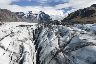İzlanda 'daki bir buzulda, arka planda karla kaplı zirveler ve parçalı bulutlu bir gökyüzü üzerine kurulmuş derin yarıklar ve volkanik kül çizgileri bulunur..