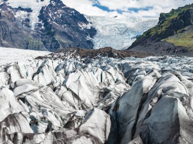 İzlanda 'daki bir buzulda parçalı buz sırtları, geniş bir buz tabakası ve parçalı bulutlu bir gökyüzünün altında yeşil bitki örtüsü bulunan karanlık volkanik dağlar bulunur..
