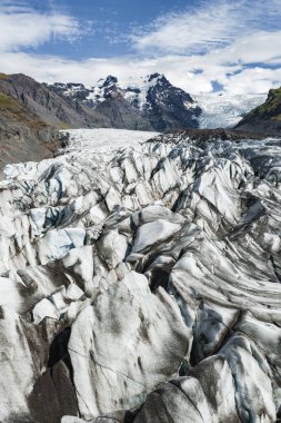Derin yarıklar ve karanlık çizgilerle İzlanda 'da engebeli karlı dağlara, kayalık araziye ve yeşil bitki örtülerine karşı kurulmuş bir buzul..