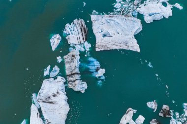 Siyah volkanik çizgileri olan büyük buzdağları turkuaz sularda yüzer. Daha küçük buz parçaları oluşumları çevreleyerek sakin bir manzara yaratıyor..