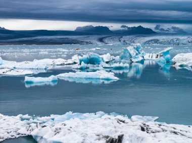 Mavi buzdağları Jokulsarlon Buzul Gölü 'nün sakin sularında yüzer, İzlanda' da bulutlu bir gökyüzünün altında bir buzulla ve engebeli tepelerle..