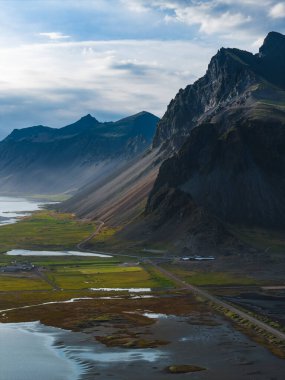 Stokknes Yarımadası 'nda dik, sivri dağlar, yansıtıcı su havuzları, yeşil alanlar ve parçalı bulutlu bir gökyüzünün altında dolambaçlı bir yol bulunur..