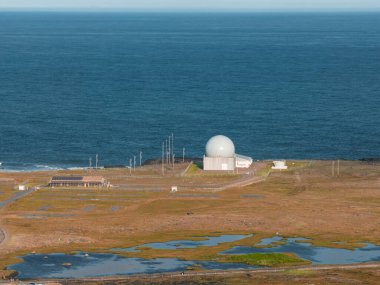 Okyanus kıyısında beyaz küresel bir radar kubbesi. Etrafı düz otlaklar, sulak alanlar, küçük su havuzları ve İzlanda 'daki görünür elektrik direkleriyle çevrili..