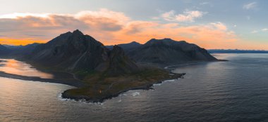 İzlanda 'nın Stokkknes Yarımadası' nın havadan görünüşü Vestrahorn 'un sivri tepeleri, siyah bir kumsal, sakin okyanus suları ve gün batımında bir deniz feneri..