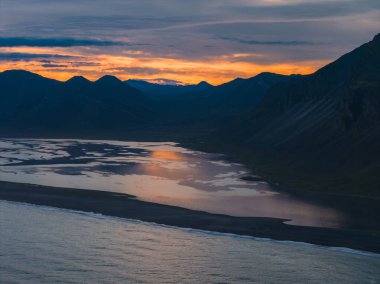 İzlanda 'nın Stokknes Yarımadası üzerinde sakin bir günbatımı, karanlık dağlar, siyah bir kumsal ve parlak turuncu gökyüzünün altında yansıtıcı su.