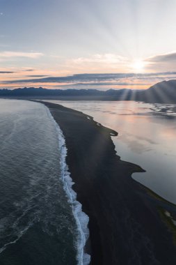 İzlanda 'da okyanus dalgaları, sakin yansıtıcı sular ve yumuşak güneş ışığı altındaki uzak bir sıradağdan oluşan siyah kumsal manzarası..