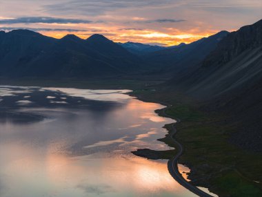Sarmal bir yol, gün batımında yeşil tarlalar ve sarp dağlarla çevrili parlak turuncu ve sarı gökyüzünün altında yansıtıcı bir su kütlesini takip eder..