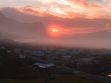 Gün batımında sakin bir İzlanda köyü, dağınık evler, dolambaçlı bir yol, alçak bir sis ve arka planda engebeli dağlar..
