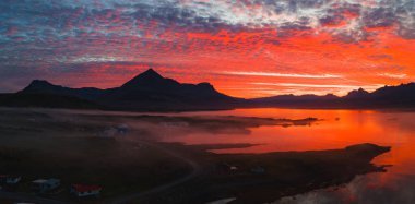 Canlı bir günbatımı, İzlanda 'da bir dağ zirvesi, kırmızı ve turuncu renkleri yansıtan sakin bir su, dağınık evler ve dolambaçlı bir yol ile aydınlatmaktadır..