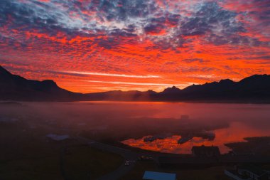 Puslu bir İzlanda kasabasının üzerinde kırmızı, turuncu ve mor renkli bir gün batımı gökyüzünü aydınlatır. Durgun su sahneyi yansıtır, sivri tepeler çerçeveler.