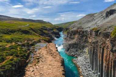 İzlanda 'daki Studlagil Kanyonu' nda bazalt sütunlar, turkuaz bir nehir, toprak kayalıklar, yeşil bitki örtüsü ve dağınık bulutlu berrak bir gökyüzü bulunur..