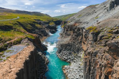 İzlanda 'daki Studlagil Kanyonu' nda dramatik bazalt sütunlar, turkuaz bir nehir, toprak kanyon duvarları, yeşil tepeler ve berrak bir gökyüzünün altında dolambaçlı bir yol bulunur..