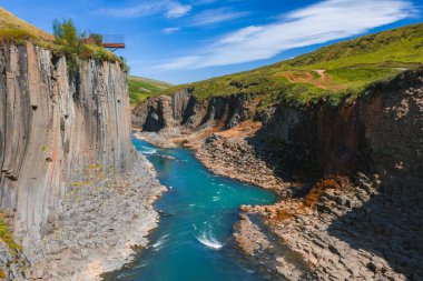 İzlanda 'daki Studlagil Kanyonu' nda bazalt sütunlar, turkuaz bir nehir, yeşil yamaçlar ve açık mavi gökyüzünün altında bir izleme platformu bulunur..