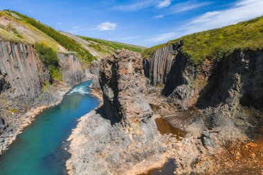 İzlanda 'daki Studlagil Kanyonu' nda etkileyici bazalt sütunlar, canlı bir turkuaz nehri, yemyeşil tepeler ve volkanik bir manzarada açık mavi bir gökyüzü bulunur..