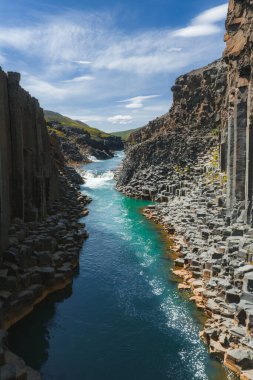 İzlanda 'daki Studlagil Kanyonu' nda keskin bazalt sütunlar, turkuaz bir nehir, küçük bir şelale ve dağınık bulutlu parlak mavi bir gökyüzü bulunur..