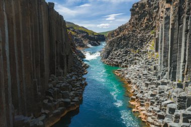 İzlanda 'daki Studlagil Kanyonu' nda yüksek bazalt sütunlar, turkuaz bir nehir, küçük bir şelale, yeşil tepeler ve parlak mavi bir gökyüzü bulunur..