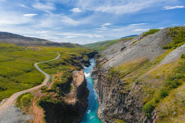 İzlanda 'daki Studlagil Kanyonu' nda dramatik bazalt sütunlar, turkuaz bir nehir, yemyeşil tepeler, kirli bir patika ve dağınık bulutlu açık mavi bir gökyüzü bulunur..