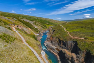 İzlanda 'daki Studlagil Kanyonu' nda bazalt sütunlar, turkuaz bir nehir, yeşil tepeler, yürüyüş yolları ve açık mavi gökyüzü altında bir izleme platformu bulunur..