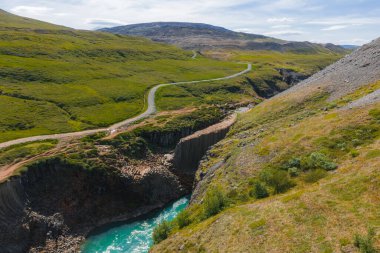İzlanda 'daki Studlagil Kanyonu' nda bazalt sütunlar, turkuaz bir nehir, yemyeşil bitki örtüsü, yuvarlanan tepeler ve solda dolambaçlı toprak bir yol bulunur..