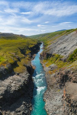 Canlı bir turkuaz nehri, açık mavi gökyüzünün altında yeşil tepeler ve engebeli arazilerle çevrili Studlagil Kanyonu 'nun bazalt kaya geçidi boyunca akar..