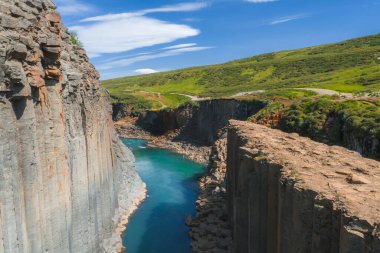 İzlanda 'daki Studlagil Kanyonu' nda geometrik bazalt sütunlar, turkuaz bir nehir, yeşil tepeler ve mavi gökyüzünün altında beyaz bulutlu bir toprak yol bulunur..