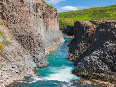 İzlanda 'daki Studlagil Kanyonu' nda yüksek bazalt sütunlar, canlı bir turkuaz nehri, yemyeşil bir çevre ve bulutlu açık mavi bir gökyüzü bulunur..