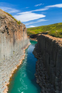 İzlanda 'daki Studlagil Kanyonu' nda bazalt sütunlar, turkuaz bir nehir ve açık mavi gökyüzünün altında dağınık bulutlarla yemyeşil bir yer alır..
