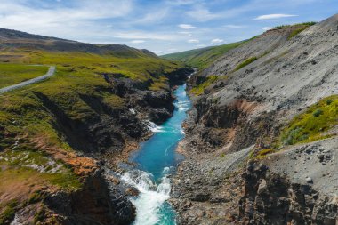Turkuaz nehri Studlagil Kanyonu 'ndan akar, bazalt oluşumları, yeşil tepeler ve dağılmış mavi bulutların altında kayalık arazi tarafından çevrelenir..
