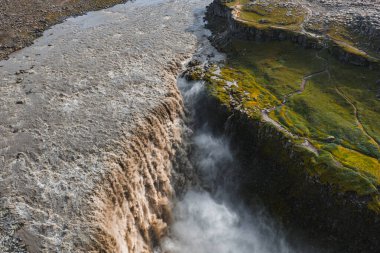 Dettifoss şelaleleri sisli bir kanyonun içine dökülür. Etrafı yosun, ot, kayalık arazi ve bir bakış açısına giden dolambaçlı bir yol ile çevrilidir..