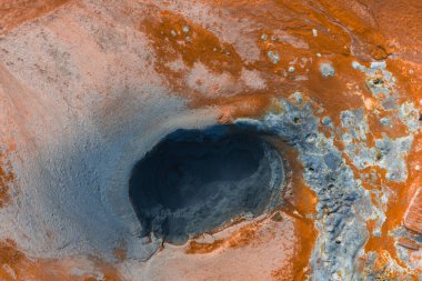 İzlanda 'daki oluşum gibi koyu renkli bir kraterin hava görüntüsü, canlı turuncu ve pas renginde arazi ile çevrili, jeotermal aktivite ve mineral yatakları gösteriyor..