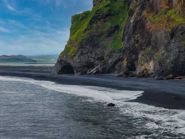 İzlanda 'da yeşil yamalı kayalıklarla çevrili siyah bir kumsal. Açılışa benzer bir mağara var. Dalgalar mavi gökyüzünün altında kıyıya vuruyor.