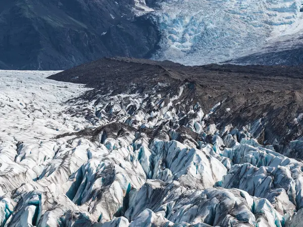 İzlanda 'daki bir buzulda derin yarıklar, kir çizgileri ve volkanik bir sırt bulunur. Arka planda mavi buz ve dağlar görülebilir..