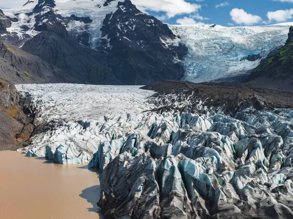Tırtıklı buz mavisi buzul oluşumları, İzlanda 'nın engebeli arazisindeki karanlık dağlar ve karla kaplı zirvelerle çerçevelenmiş çamurlu bir buzul gölüne yol açar..