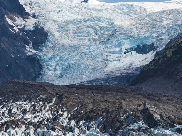 Engebeli yarıkları olan buzlu mavi buzullar karla kaplı bir dağdan iniyor. Tırtıklı buz ve volkanik enkaz kontrastı yosun kaplı bir yosunla kaplı..