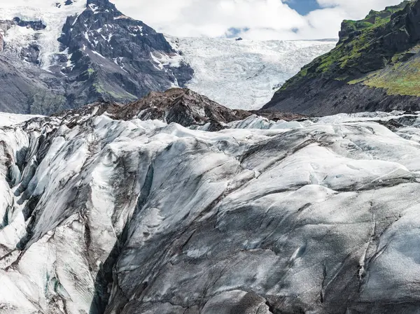 Yeşil yamalı dik volkanik dağlar ve İzlanda 'nın engebeli arazisinde parçalı bulutlu bir gökyüzü tarafından çerçevelenmiş bir buzul ön plana doğru uzanır..