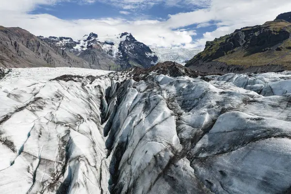 İzlanda 'daki bir buzulda, arka planda karla kaplı zirveler ve parçalı bulutlu bir gökyüzü üzerine kurulmuş derin yarıklar ve volkanik kül çizgileri bulunur..