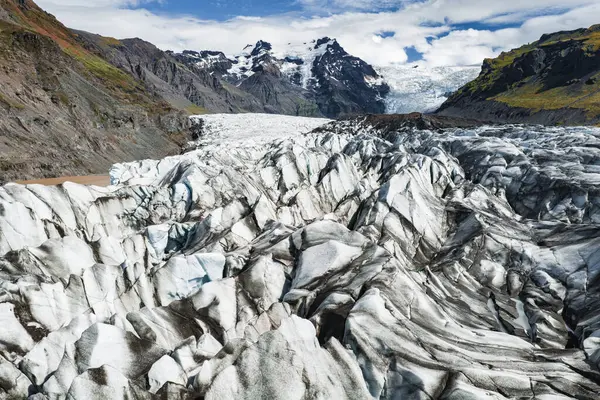 Bir buzulun sivri buz yüzeyi ufka doğru uzanır, yosunlu yosunlar ve yosunlu yamaçlarla çevrili, bulutlu bir gökyüzünün altında karla kaplı dağlar..
