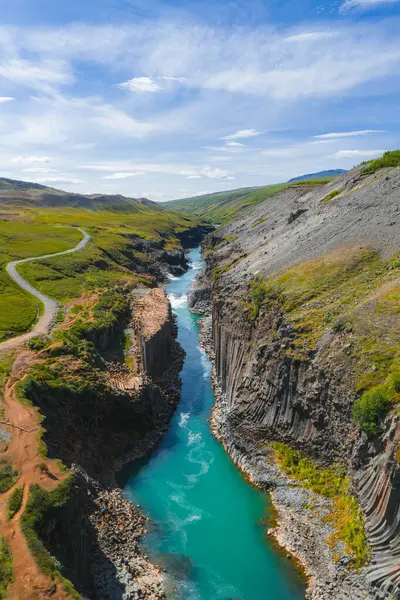 Studlagil Kanyonu bazalt kolonları, turkuaz nehri, yeşillik, kayalık arazi, toprak yol, dolambaçlı yol ve bulutlu açık mavi gökyüzü içerir..