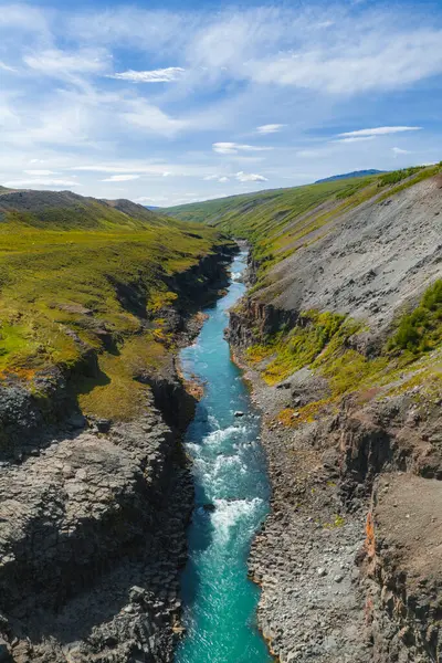 Canlı bir turkuaz nehri, açık mavi gökyüzünün altında yeşil tepeler ve engebeli arazilerle çevrili Studlagil Kanyonu 'nun bazalt kaya geçidi boyunca akar..