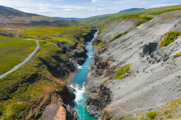 İzlanda 'daki Studlagil Kanyonu' nda turkuaz bir nehir, dik kayalık uçurumlar, yeşil bitki örtüsü, toprak yol ve uzak tepeleri olan açık mavi bir gökyüzü bulunur..