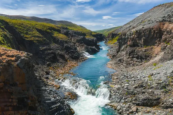 İzlanda 'daki Studlagil Kanyonu' nda parlak mavi bir nehir, engebeli bazalt sütunlar ve açık mavi gökyüzü altında yeşil bir bitki örtüsü bulunur..