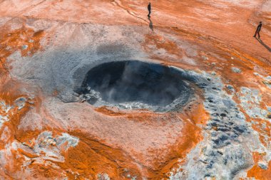 İzlanda 'da buharlaşan bir jeotermal kaplıcanın havadan görünüşü, turuncu ve beyaz mineral yataklarıyla çevrili. Yakınlardaki kırmızı arazide iki kişi duruyor..