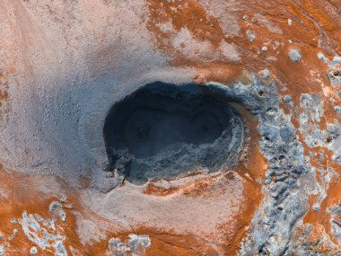 İzlanda 'da, turuncu ve gri mineral yataklarıyla çevrili kaynayan bir jeotermal çamur tenceresinin hava görüntüsü. Arazi canlı jeotermal aktiviteler sergiliyor.