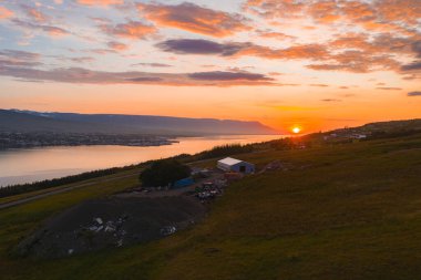 Gün batımında sakin bir İzlanda fiyordu, uzak dağlarla ve küçük bir kasabayla sınırlıydı. Ön planda çimenli bir yamaçta dağınık yapılar var..