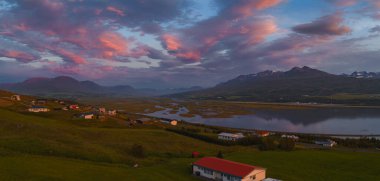 Pembe ve mor renkli canlı bir günbatımı küçük bir İzlanda köyünü aydınlatır, yemyeşil alanlar, sakin sular, dolambaçlı dereler ve uzak dağlar..