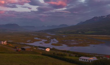 Canlı pembe ve mor bir gün batımı, İzlanda 'nın verimli bir vadisinde, karlı dağlarla çevrili ve dağınık kırsal evlerle çevrili dolambaçlı bir nehri yansıtır..