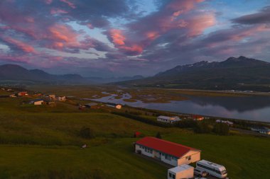 Gün batımında İzlanda kırsalının sakin hava manzarası. Sakin bir nehir, ön planda evler ve yeşil tarlalarla pembe ve turuncu renkleri yansıtır..